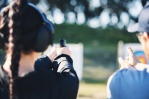 Woman, gun and learning to shoot outdoor with instructor at shooting range for target training. Safety and security with hand teaching person sport game or aim with gear and firearm for focus
